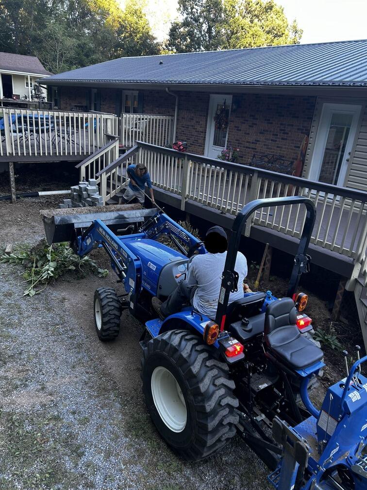 2 Workers are using a rake and tractor to landscape around a deck.